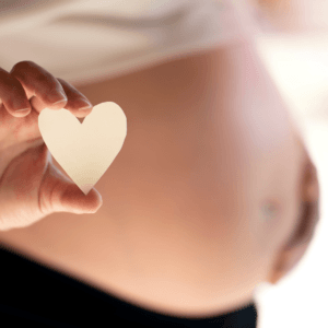 Ventre rond de femme enceinte. La femme touche son ventre et avec son autre main tient un coeur blanc - Stéphanie Périnatalité